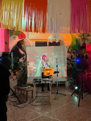 Una performer con una parrucca rosa davanti a una proiezione, sotto una tenda di frange colorate.